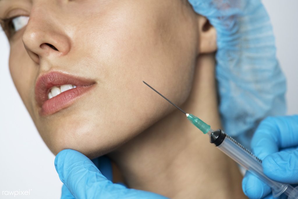 White female patient with blue hair cap on holds still as a needle is held up to her face for the non-surgical face lift.